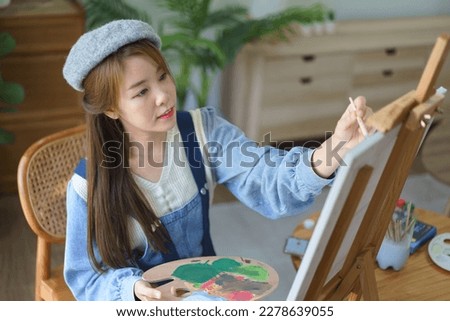 Young female painter holds color palette and use brush to painting artwork on canvas in home studio.