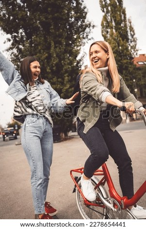 Vertical photo of two young women having fun in the city, one riding a red bike while other smiles with hands in the air behind her 