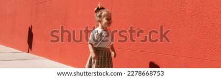 happy toddler girl in checkered skirt and white t-shirt walking near building with red wall, banner