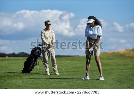 Full length portrait of sporty young couple playing golf in green field with focus on black woman hitting ball with club Royalty-Free Stock Photo #2278669983