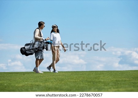 Minimal side view portrait of elegant sporty couple carrying golf bag walking on green field against blue sky, copy space Royalty-Free Stock Photo #2278670057
