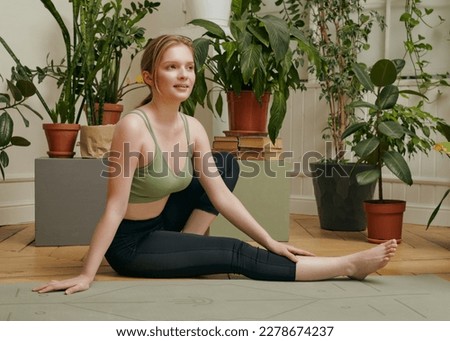 a happy woman is sitting at home surrounded by green plants resting after a workout