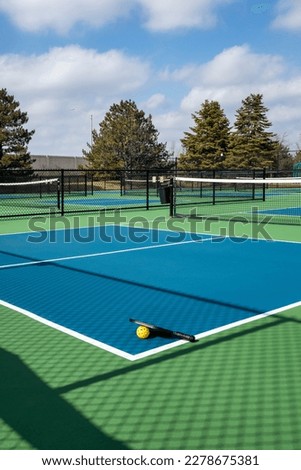 View of a pickleball complex with a paddle and yellow ball on blue and green courts beside a playground in a suburban park in early spring. Royalty-Free Stock Photo #2278675381