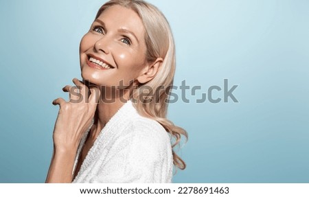 Smiling female aging model, clear glowing skin, nourished face without blemishes, applies daily cream, hyaluron anti-aging treatment, looking in mirror at herself, blue background. Royalty-Free Stock Photo #2278691463