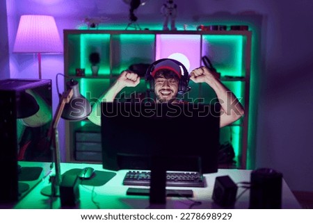 Young hispanic man streamer using computer and headphones with winner expression at gamin room
