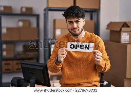 Hispanic man with beard working at small business ecommerce holding open sign clueless and confused expression. doubt concept. 