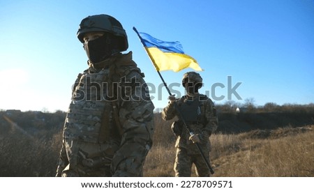 Young woman and man in military uniform going with national banner at meadow. Female and male soldier of ukrainian army walking with flag of Ukraine at field. Victory against russian aggression.