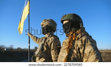 Young soldiers of ukrainian army standing at peak of hill with raised national banner. Woman and man in camouflage uniform looking at distance with lifted flag of Ukraine against blue sky. Close up.