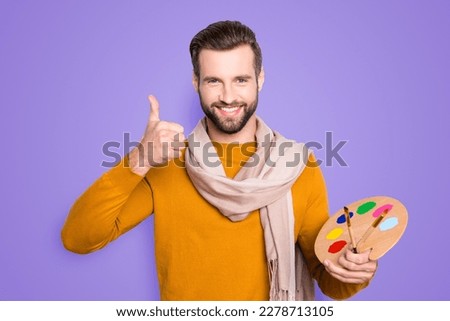Portrait of stylish cheerful artist with hairstyle, stubble in pullover and scarf around his neck showing thumbup with finger holding colorful palette and brushed in hand over grey background