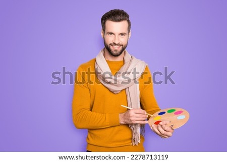 Portrait of positive attractive artist with stubble, hairstyle in sweater and scarf on neck using having colorful palette and brushed in arms, isolated on grey background