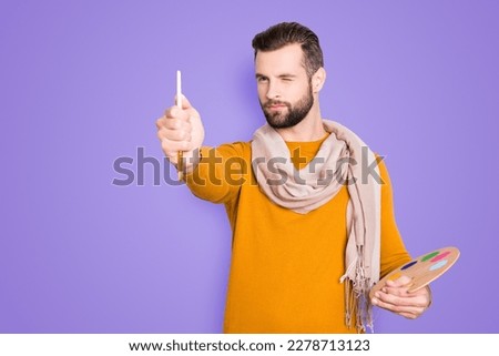 Portrait of creative concentrated artist with bristle in sweater and scarf on neck using, having colorful palette, brushed in arms, analyzing, expertising pic, isolated on grey background