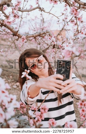 Vertical picture of a pretty smiling woman with sunglasses and a hipster jumper taking a selfie portrait in a cherry blossom field during springtime. Sunset time and soft light. Social media concept