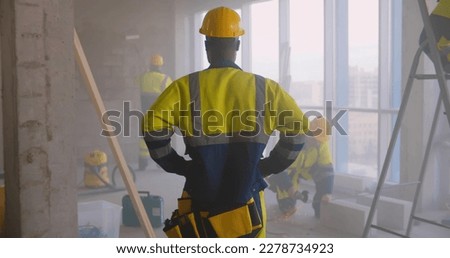 Back view of African-American foreman in uniform supervising team of builders at construction site. Black constructor in hardhat watching his colleagues renovating apartment