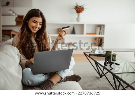 Smiling millennial mixed race woman shopaholic typing on laptop, using credit card, enjoy online shopping in living room interior. Business remotely, sale, banking and cashback at home