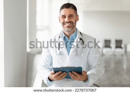 Professional help. Portrait of happy male general practitioner standing with clipboard and smiling at his office in clinic. Doctor wearing stethoscope and white coat Royalty-Free Stock Photo #2278737007