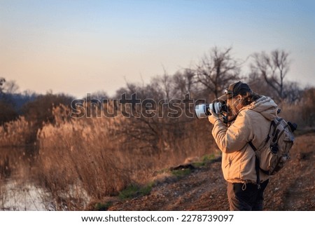 Landscape photographer with camera and telephoto lens photographing wildlife in nature during sunset