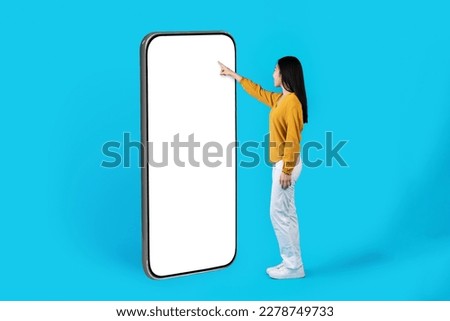 Young slim asian woman in casual outfit touching white blank mockup screen of big smartphone, choosing goods on webstore, scrolling on social media, websurfing, blue studio background, copy space Royalty-Free Stock Photo #2278749733
