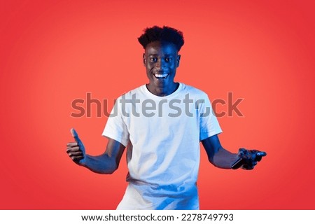 Happy handsome young black guy in white winner champion holding joystic and showing thumb up, isolated on red background in neon light, modern technologies in entertainment, video games