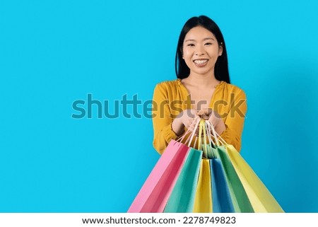 Positive cute pretty young asian woman in yellow holding colorful shopping bags and smiling at camera, isolated on blue studio background, copy space. Season sale, black friday concept