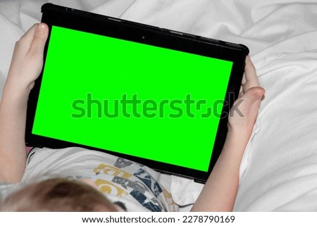 Top view a little girl in a white bed is holding a tablet with a green chroma key screen. Watching a cartoon or an educational lesson