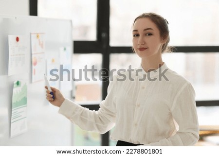 Young lady manager in white blouse points to statistics papers on whiteboard looking in camera. Woman conducts business seminar in office