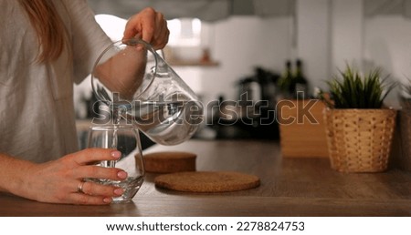 Faceless young woman pour pure water from jug to glass in kitchen. Anonymous girl in white t-shirt holds jar takes care of water balance. High quality photo Royalty-Free Stock Photo #2278824753