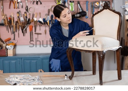 To replace upholstery, young female restorer carefully removes old cloth seat covering from antique wooden chair. Royalty-Free Stock Photo #2278828017
