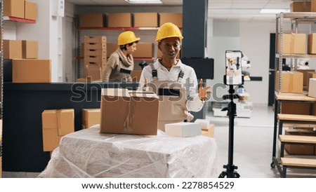 African american woman advertising products in boxes working with smartphone in storage room. Female employee in overalls filming advertisement video for social media, industrial stock.