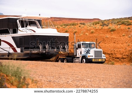 Big powerful American bonnet rig semi truck with sign oversize load transporting down the red rocks hill to the water a huge oversize multilevel ship on marine resort at Lake Powell in Page Arizona Royalty-Free Stock Photo #2278874255