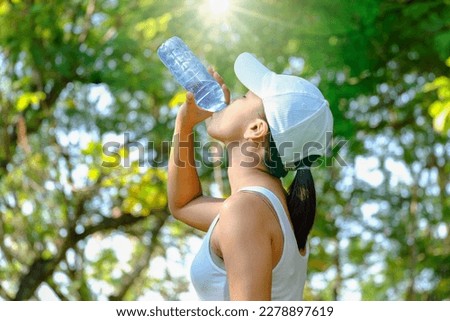 Female athlete lifts a bottle of drinking water in a clean bottle in hot weather with very sunny natural green background. Health and hydration concepts Royalty-Free Stock Photo #2278897619