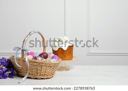 Easter basket with painted eggs near tasty cake and bouquet of flowers on white marble table. Space for text