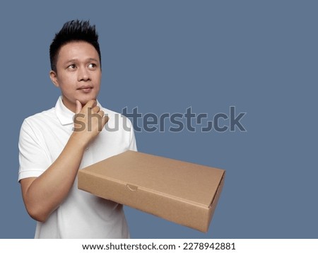 Man holding cardboard box and finger holding chin isolated on plain background.