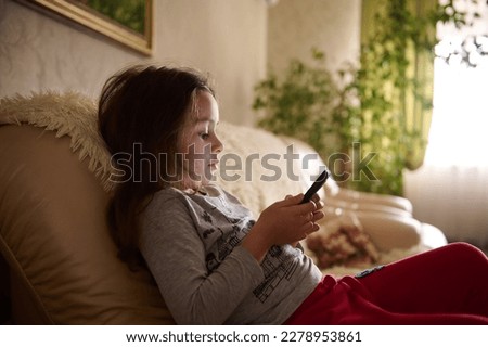 Cute little child girl watching online movie or video, playing virtual game in mobile phone while sitting on couch at home. The problem of gadget addiction in childhood. Kids and digital technologies