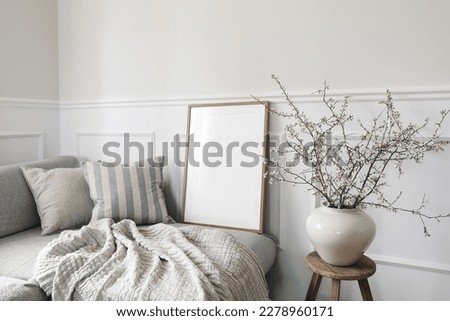 Modern spring scandinavian living room interior. Wooden picture frame, poster mockup. Sofa with linen pale blue striped cushions. Cherry plum blossoms in vase. Elegant stylish minimal home decor.