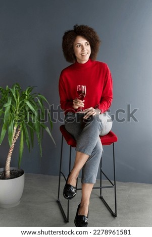 Full-length portrait attractive confident elegant multi-ethnic woman with afro hair, sitting on a chair in a modern interior, holding a wine glass and taking a sip of red wine, pensively looking away