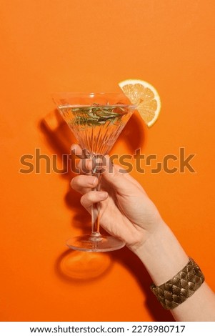 Woman holding martini glass of refreshing cocktail with lemon slice and rosemary on orange background, closeup