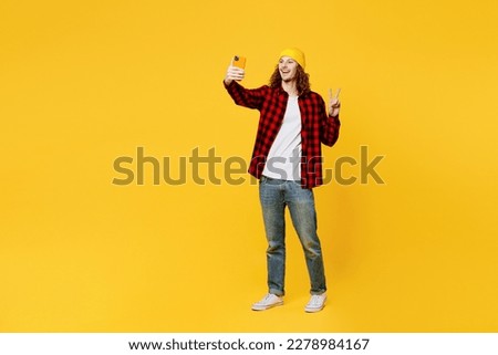 Full body young man wearing red checkered shirt white t-shirt hat doing selfie shot on mobile cell phone post photo on social network show v-sign isolated on plain yellow background studio portrait