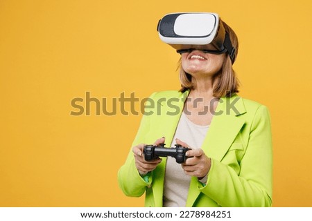 Elderly gambling fun woman 50s years old wear green jacket white t-shirt hold play pc game with joystick console watching in vr headset pc gadget isolated on plain yellow background studio portrait
