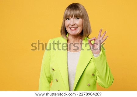 Elderly smiling happy satisfied woman 50s years old wearing green jacket white t-shirt showing okay ok gesture look camera isolated on plain yellow background studio portrait. People lifestyle concept