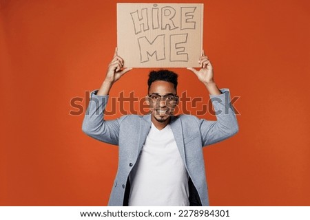Young fun employee business man corporate lawyer wear classic formal grey suit shirt glasses work in office hold cardboard sign card hire me above head isolated on plain red orange background studio