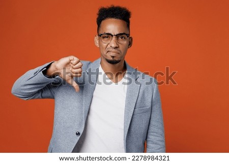 Young sad employee business man corporate lawyer wear classic formal grey suit shirt glasses work in office showing thumb down dislike gesture distrust isolated on plain red orange background studio