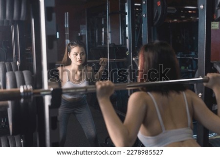 A young and fit woman does barbell squats in front of the mirror at the gym. Using the bar weight only. Royalty-Free Stock Photo #2278985527