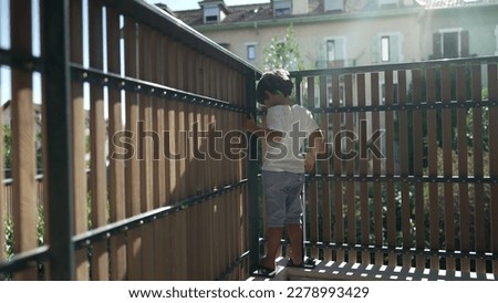 One playful child standing at apartment balcony looking through wooden fence protection from second floor of residential home