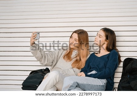 Two teenage girls taking selfie on the phone while sitting on a bench near the university. People, friendship, studying, lifestyle concept	                             