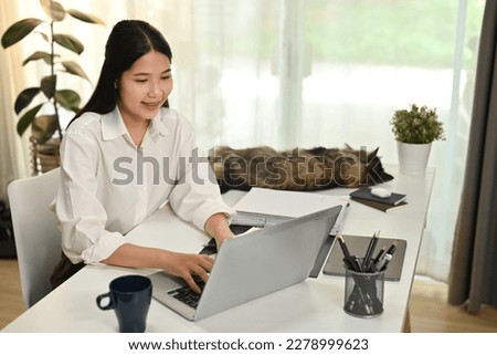 Happy young female freelancer sitting at home office desk with cat and working online on laptop computer