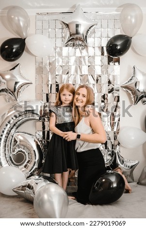 Mom congratulate daughter on her birthday near arch. Happy family celebrating a birthday party 6 years. Mother hugging child. Photo wall decoration white, black, silver balloons. Funny time together.