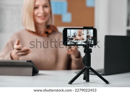 Close-up of smartphone standing on tripod with teacher recording online lesson for her students at table in class Royalty-Free Stock Photo #2279022453