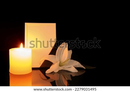 Blank funeral frame, lily flower and burning candle on dark background