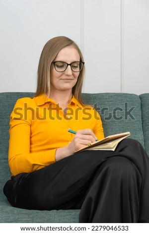 Woman in an orange shirt, black trousers and glasses is sitting on the couch taking notes in a notebook. At a psychologist's appointment. Business, journalist, psychologist, coach
