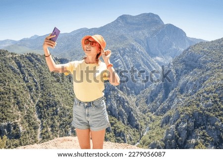 Girl taking selfie photos on her smartphone from a scenic viewpoint with majestic panorama of mountain canyon
